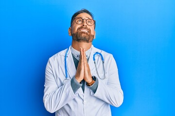 Handsome middle age man wearing doctor uniform and stethoscope begging and praying with hands together with hope expression on face very emotional and worried. begging.