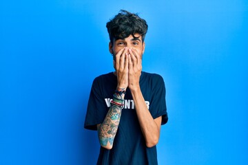 Young hispanic man wearing volunteer t shirt laughing and embarrassed giggle covering mouth with...
