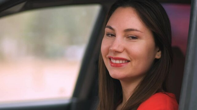 A Young Beautiful Woman In A Red Dress In A Red Car Rolls Down The Window And Smiles. Girl Driver On The Road