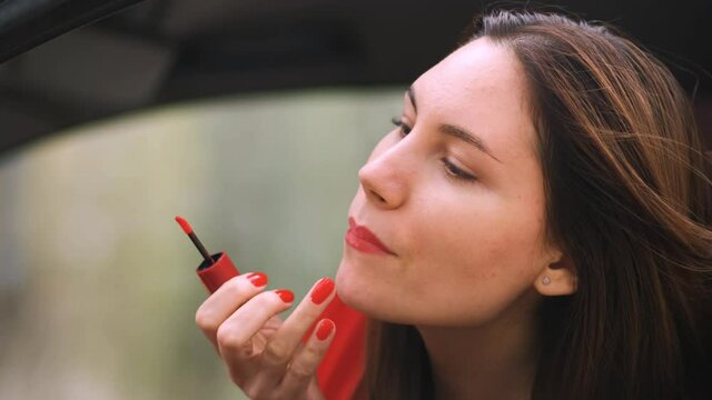 A young woman in a red dress in a red car looks in the mirror and paints her lips red. Girl driver preening on the road. Red lipstick