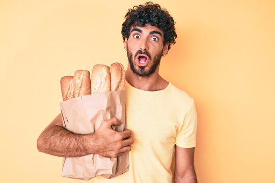 Handsome young man with curly hair and bear holding paper bag with bread scared and amazed with open mouth for surprise, disbelief face