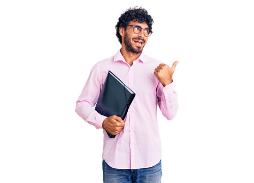 Handsome young man with curly hair and bear holding business folder pointing thumb up to the side smiling happy with open mouth