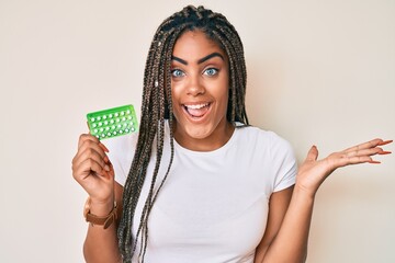 Young african american woman with braids holding birth control pills celebrating victory with happy...