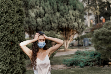 Headache girl in blue medical mask. Woman touches her head because sicks coronavirus covid-19. Pandemic 2020. Attractive girl weared white formal dress in green park