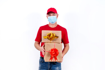 Safe contactless remote delivery of holiday gifts during the coronavirus pandemic. A courier in a red uniform and protective medical mask holds two beautiful gift boxes with bows