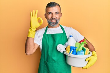 Middle age handsome man holding cleaning products doing ok sign with fingers, smiling friendly gesturing excellent symbol