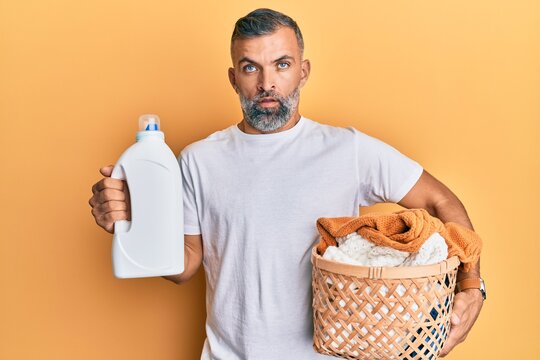 Middle age handsome man holding laundry basket and detergent bottle in shock face, looking skeptical and sarcastic, surprised with open mouth