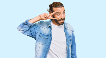 Fototapeta premium Attractive man with long hair and beard wearing casual denim jacket doing peace symbol with fingers over face, smiling cheerful showing victory