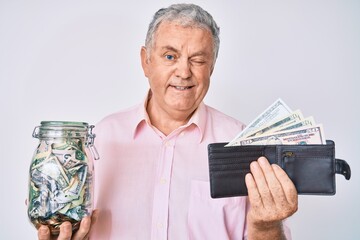Senior grey-haired man holding wallet with dollars and jar with savings winking looking at the...