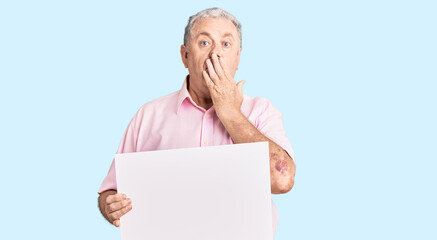 Senior grey-haired man holding blank empty banner covering mouth with hand, shocked and afraid for mistake. surprised expression