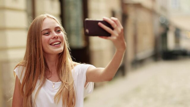 Young smiling hipster blond woman in white t-shirt posing, takes selfies and streams video to social media from her smartphone.