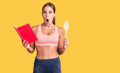 Young beautiful hispanic woman wearing gym clothes reading cooking recipe book holding spoon scared and amazed with open mouth for surprise, disbelief face