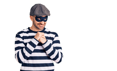 Young handsome man wearing burglar mask laughing nervous and excited with hands on chin looking to the side