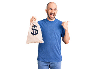 Young handsome man holding money bag with dollar symbol pointing thumb up to the side smiling happy with open mouth
