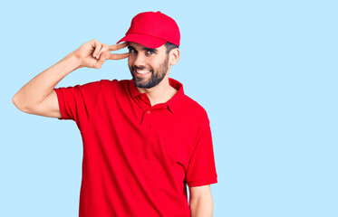 Young handsome man with beard wearing delivery uniform doing peace symbol with fingers over face, smiling cheerful showing victory