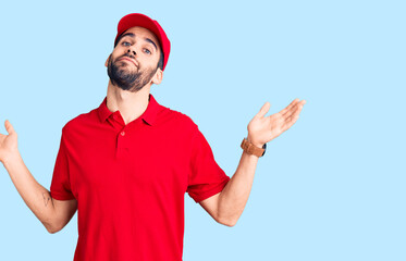 Fototapeta premium Young handsome man with beard wearing delivery uniform clueless and confused expression with arms and hands raised. doubt concept.