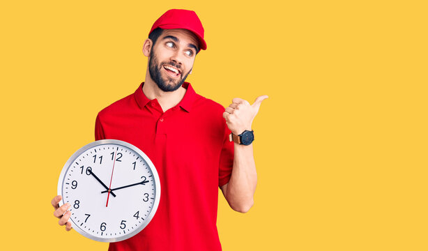 Young handsome man with beard wearing delivery uniform holding big clock pointing thumb up to the side smiling happy with open mouth