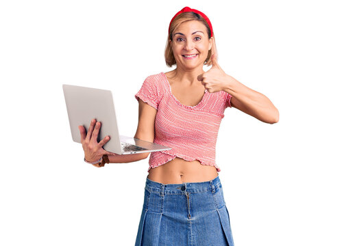 Young blonde woman holding laptop smiling happy and positive, thumb up doing excellent and approval sign