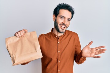Young hispanic man holding take away food celebrating achievement with happy smile and winner expression with raised hand