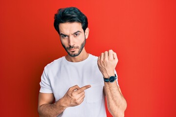 Young hispanic man wearing casual white tshirt in hurry pointing to watch time, impatience, looking...