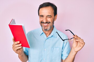 Middle age hispanic man reading book holding glasses winking looking at the camera with sexy...