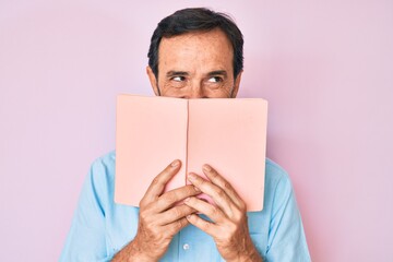 Middle age hispanic man reading book smiling looking to the side and staring away thinking.