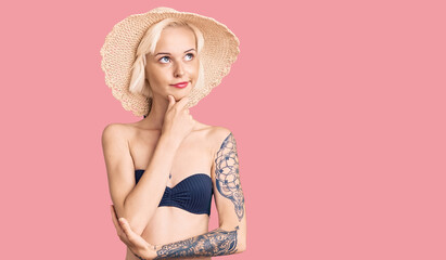 Young blonde woman with tattoo wearing bikini and summer hat with hand on chin thinking about question, pensive expression. smiling with thoughtful face. doubt concept.