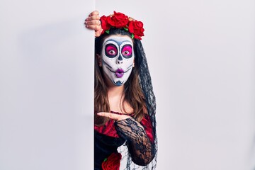 Young woman wearing day of the dead custome holding blank empty banner surprised pointing with finger to the side, open mouth amazed expression.