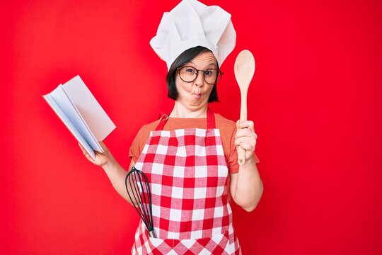 Brunette woman with down syndrome wearing professional baker apron reading cooking recipe book making fish face with mouth and squinting eyes, crazy and comical.