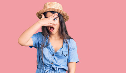 Brunette teenager girl wearing summer hat peeking in shock covering face and eyes with hand, looking through fingers with embarrassed expression.