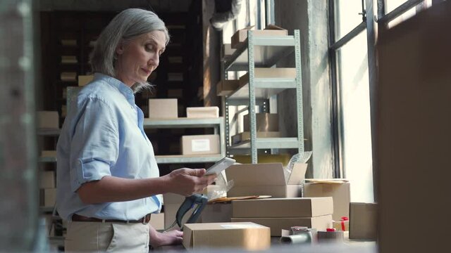 Female mature senior small business owner using mobile app checking parcel box. Warehouse worker, seller holding phone scanning retail dropshipping package postal parcel on cell preparing ship order.