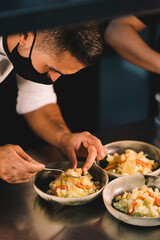 Close-up of a male chef with mask decorating food in ceramic dishes over stainless steel worktop in restaurant kitchen during coronavirus pandemic.
