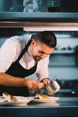 Foto op Plexiglas Portrait of a male chef decorating food with spoon in ceramic dish over stainless steel worktop in restaurant kitchen. © Henko Studio