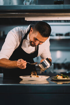 Portrait of a male chef decorating food with spoon in ceramic dish over stainless steel worktop in restaurant kitchen.