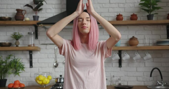 Smiling transwoman with pink wig and t-shirt does namaste practicing yoga in contemporary decorated kitchen with new furniture