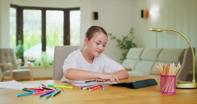 Focused teen girl, at the desk, scrolls tablet, then stretches raising hands. Felt-tip pens, white paper, tablet, pencils, books lamp on the desk. Walls, window on background are blurred