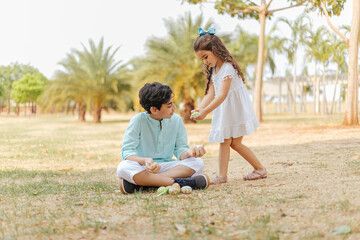 Latin brothers celebrating Easter. Boy sitting on the grass in a park and curly girl holding easter eggs on a sunny day