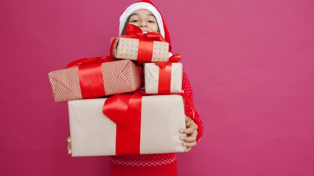 Funny indian latin preteen girl wears santa hat holding many gifts dropping boxes falling down standing on red background. Merry Christmas presents shopping sale, Happy 2021 New Year celebration.