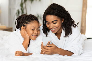 Obraz na płótnie Canvas Little black girl and her mom listening music on smartphone, wearing bathrobes