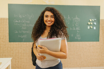 Latin Curly haired student smiling wearing backpack holding a notebook in a classroom - obrazy, fototapety, plakaty