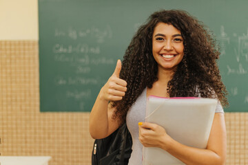 Latin Curly haired student smiling wearing backpack holding a notebook in a classroom with Thumbs up