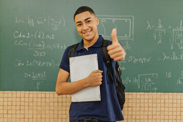 Latin men student smiling wearing backpack holding a notebook in a classroom with Thumbs up