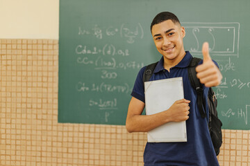 Latin men student smiling wearing backpack holding a notebook in a classroom with Thumbs up