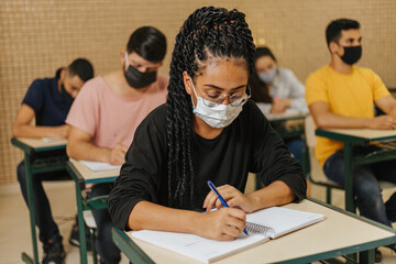 Latin students in the classroom. female student with twisted hairstyle and wearing mask writing in notebook with a pen
