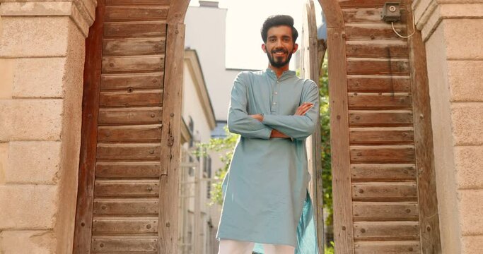 Portrait shot of Hindu handsome young man in traditional outfit standing outdoor at street in wooden door and smiling to camera. Attractive cheerful male model in India.