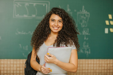 Fototapeta Latin Curly haired student smiling wearing backpack holding a notebook in a classroom obraz