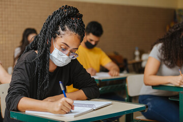 Latin students in the classroom. female student with twisted hair wearing mask and writing in...