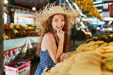 A young smilling  redhead girl in hat holding near yellow mango in her hand at the marketplace in Thailand. Copy space