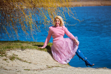 Attractive blonde woman sits under a tree, beside lake wearing a classic pink wool polo neck...
