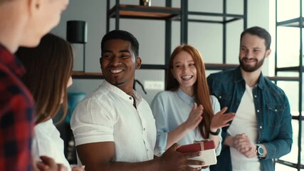Happy team of multi-ethnic business people congratulate African-American colleague and give gift with surprise. Cheerful birthday man is surprised to open the box with gift to applause of coworkers.